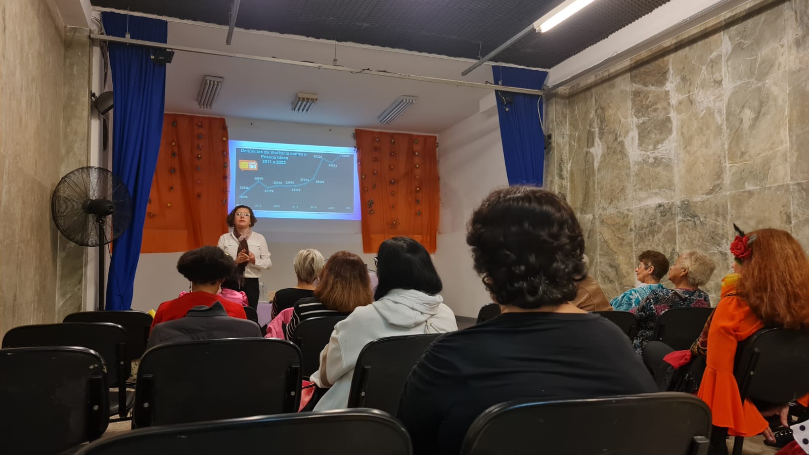 10 pessoas se sentam em cadeiras pretas, viaradas para frente. Uma mulher branca de cabelos cacheados, vestindo blusa branca e calça preta, está na frente de todos falando. Ao lado dela, sob um palco cinza, há uma mesa coberta por toalha roxa com uma jarra de água em cima e outra mesa ao lado, também coberta com toalha roxa. Em cima da mesa, há um projetor preto que projeta uma apresentação de PowerPoint na parede branca. Em volta da apresentação há uma cortina laranja. Dos dois lados do palco há ventiladores pretos e uma cortina azul.